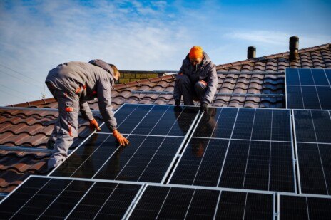 Two individuals working on solar panels on roof of building. One person bending over, wearing gray pants with orange stripe down side, red hat, and black gloves. Another person kneeling beside them, wearing gray jacket, white pants, and black gloves. Solar panels are dark blue with silver edges and are arranged in rows on roof of building. The sky is clear and blue above the scene.